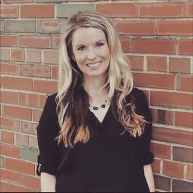 A smiling light-skinned female with long blonde hair wearing a black shirt against a brick wall backdrop