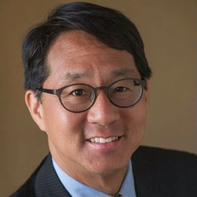 A smiling man of Asian heritage with short black hair wearing glasses and a suit and tie against a brown background