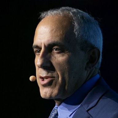 An image of an older adult man with white and slightly tan skin and short white hair. It is an image of mostly his head and he is speaking into an earpiece microphone and wearing a blue suit and tie. He is against a black background. 