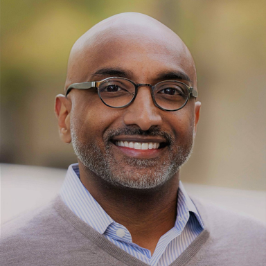 An image of a bald adult man with a stubbly beard and brown skin. He is wearing round glasses and a gray sweater and smiling at the camera.