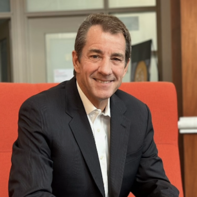 person with short cropped hair in black suit and white button up shirt sitting in red chair