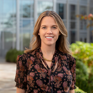 A smiling light-skinned female-presenting person named Agnes Stephens with light brown hair wearing a pink and black patterned shirt against an outdoor background.