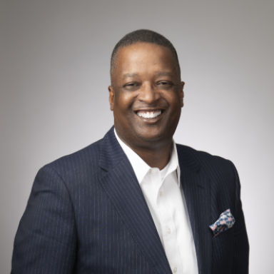 An image of an adult man with brown skin and short dark hair. He is smiling at the camera and wearing a navy blue blazer over a dress shirt against a grey background.