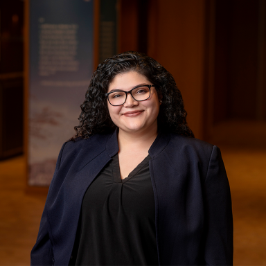 A smiling tan-skinned woman named Glorimar Aragon with long black hair, wearing a black blazer with a black shirt and glasses, and standing in a room with dark wood details.