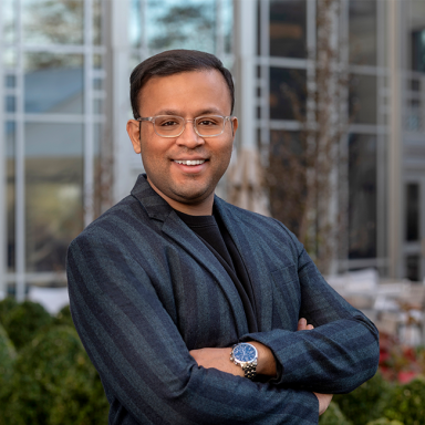 An image of a brown skinned, smiling, adult man named Ishtiaq Mawla with his arms crossed. He has short dark hair and is wearing glasses and a dark blue striped suit. 