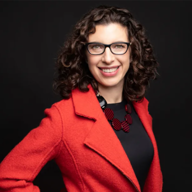 A photo of a women with light white skin against a black background. She has dark shoulder-length curly hair, and she is wearing glasses and a red blazer while smiling at the camera.