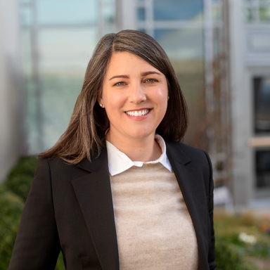 An image of a female-presenting person named Nadia Penrod. She has medium length brown hair and white skin. She is smiling at the camera and wearing a black jacket with a tan sweater. 