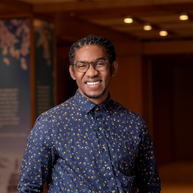 A smiling dark-skinned man named Quinton Banks with short dark braided hair, wearing glasses and a navy blue patterned dress shirt, and standing in a room with dark wood details.