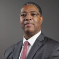 A darker-skinned male with gray hair wearing glasses and a gray suit and tie against a gray backdrop