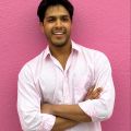 A smiling darker-skinned male wearing a light pink button-up shirt against a pink background