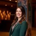 A smiling light-skinned woman named Emily Musil with long brunette hair, wearing a green blazer, and standing in a room with dark wood details.