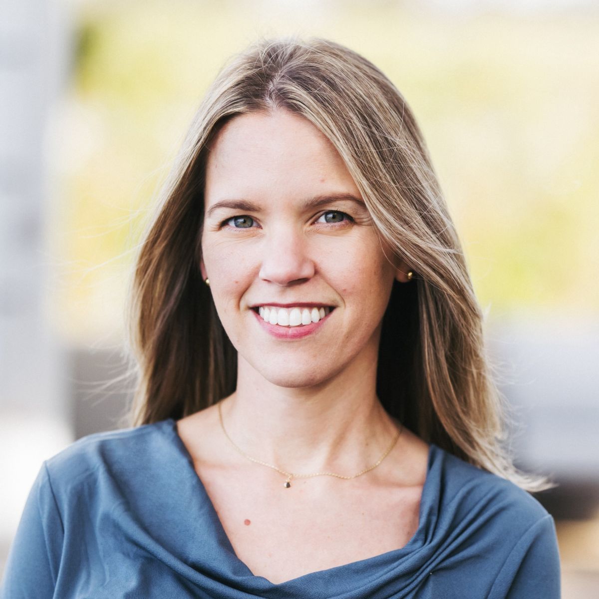 A smiling light-skinned female-presenting person with light brown hair wearing a blue shirt against an outdoor background