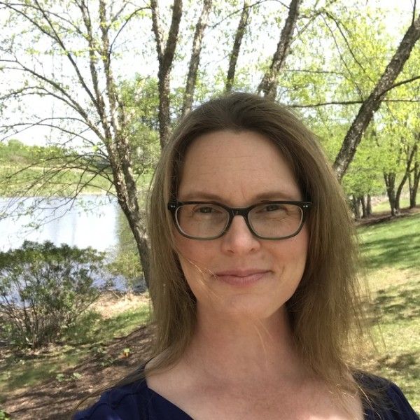 A smiling light-skinned female-presenting person with light hair against an outdoor background.