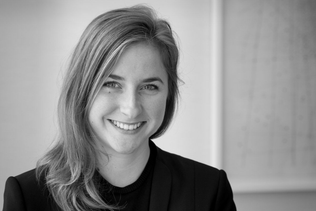 Smiling light-skinned, female-presenting individual with long length blonde hair wearing a black shirt on a grey backdrop.