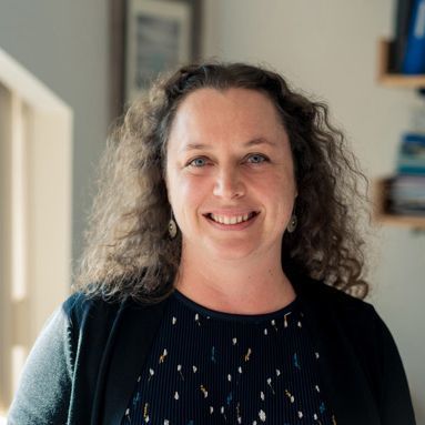 A smiling light-skinned female-presenting person with dark curly hair wearing a black shirt and jacket against an indoor background