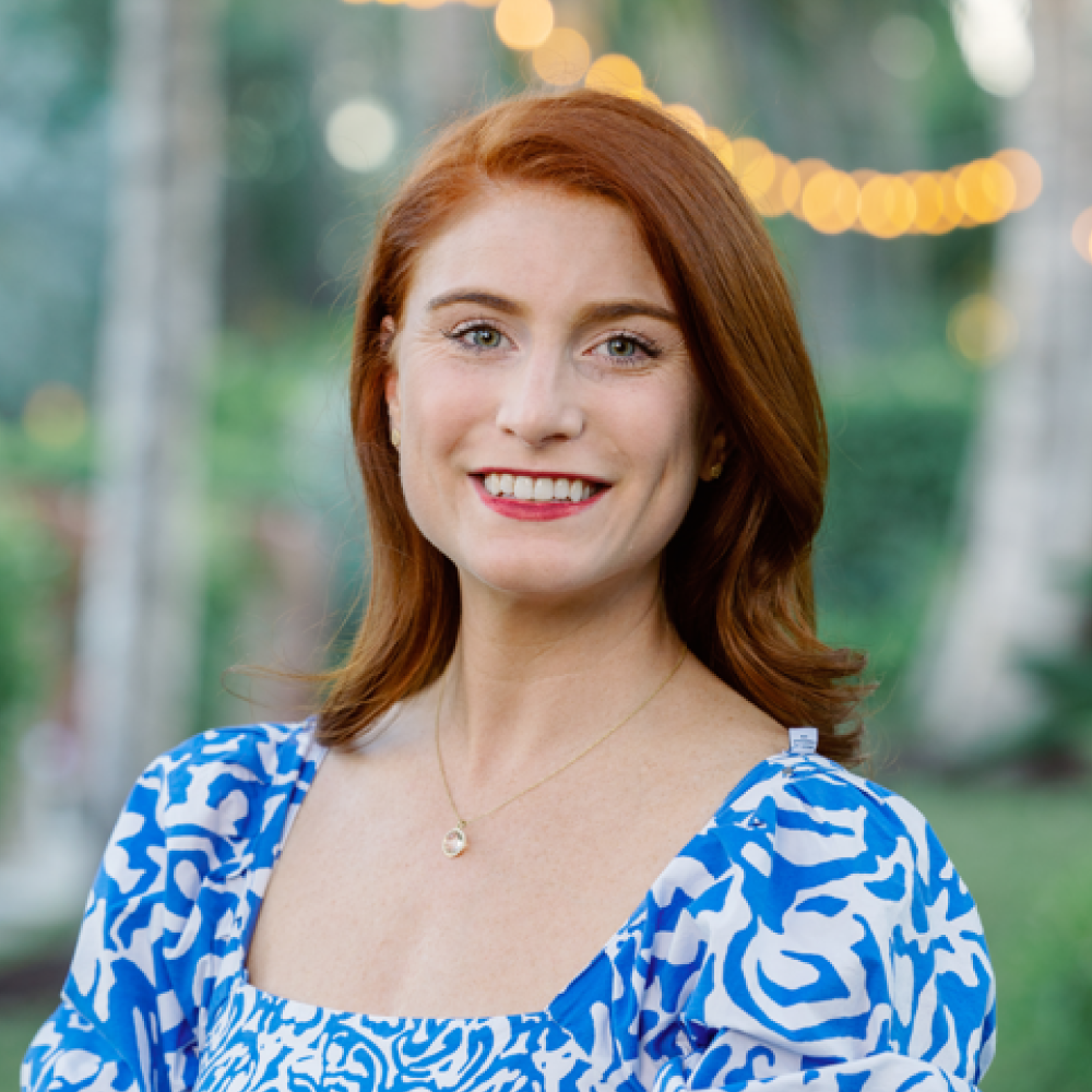 Smiling light-skinned female-presenting person with red hair wearing a blue and white patterned shirt in front of an outdoor background