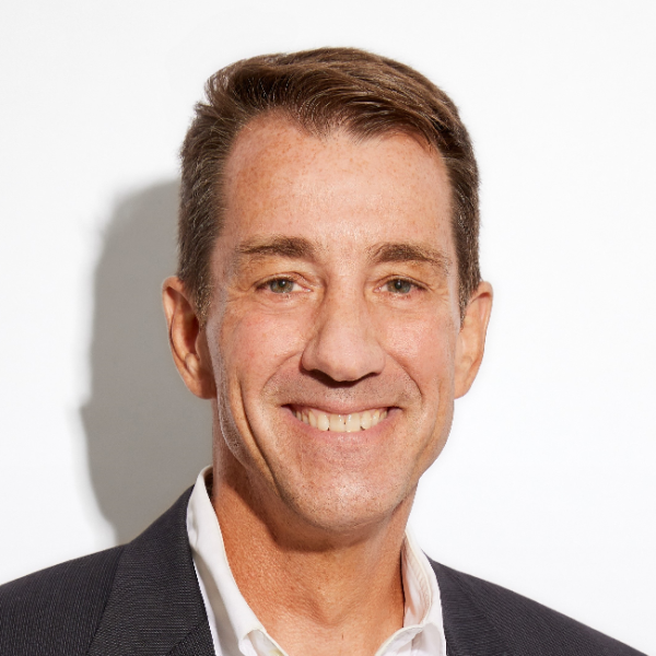 A smiling light-skinned male-presenting person with short, brown hair wearing a suit jacket against a white background