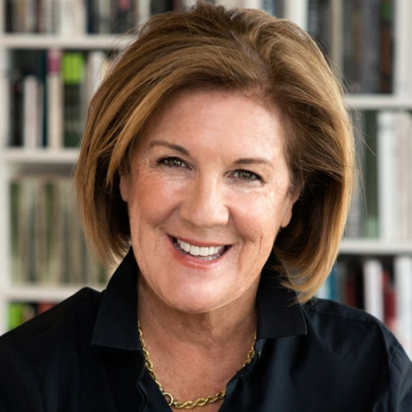 A smiling female-presenting person with brown hair wearing a black top against a blurred background of books.