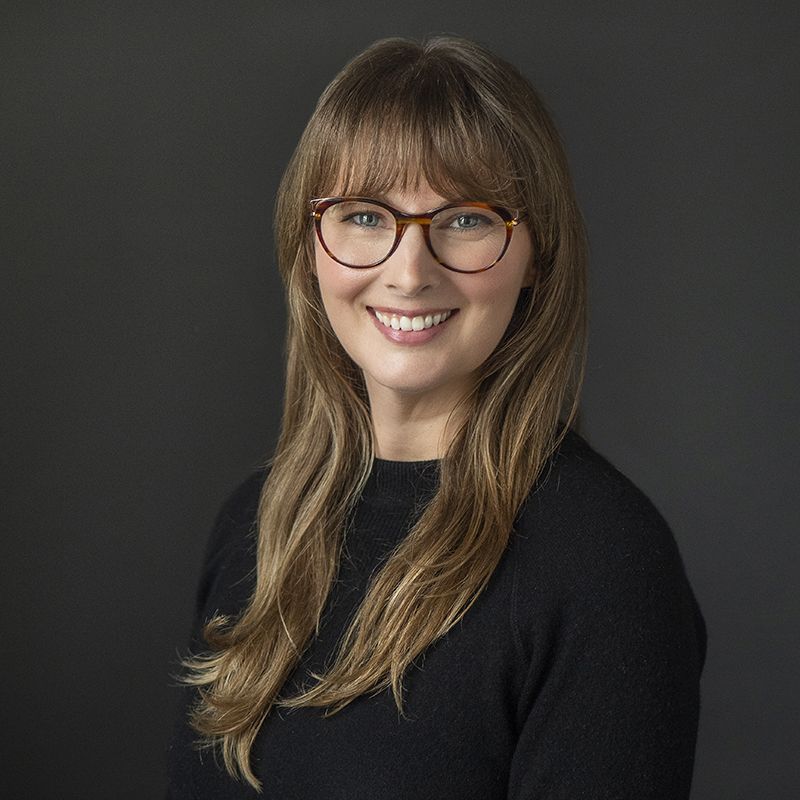 Smiling light-skinned, female-presenting individual with long length blonde hair wearing a black shirt on a grey backdrop.