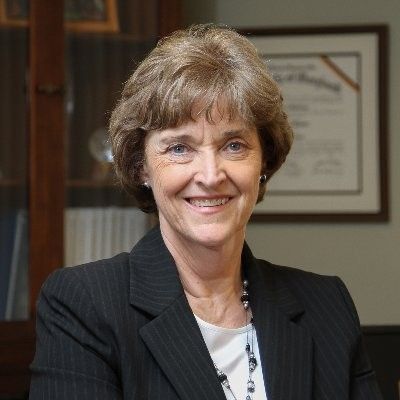 A smiling light-skinned, female-presenting person with brown hair wearing a dark suit jacket and white shirt against a background containing a bookshelf and academic degree frame