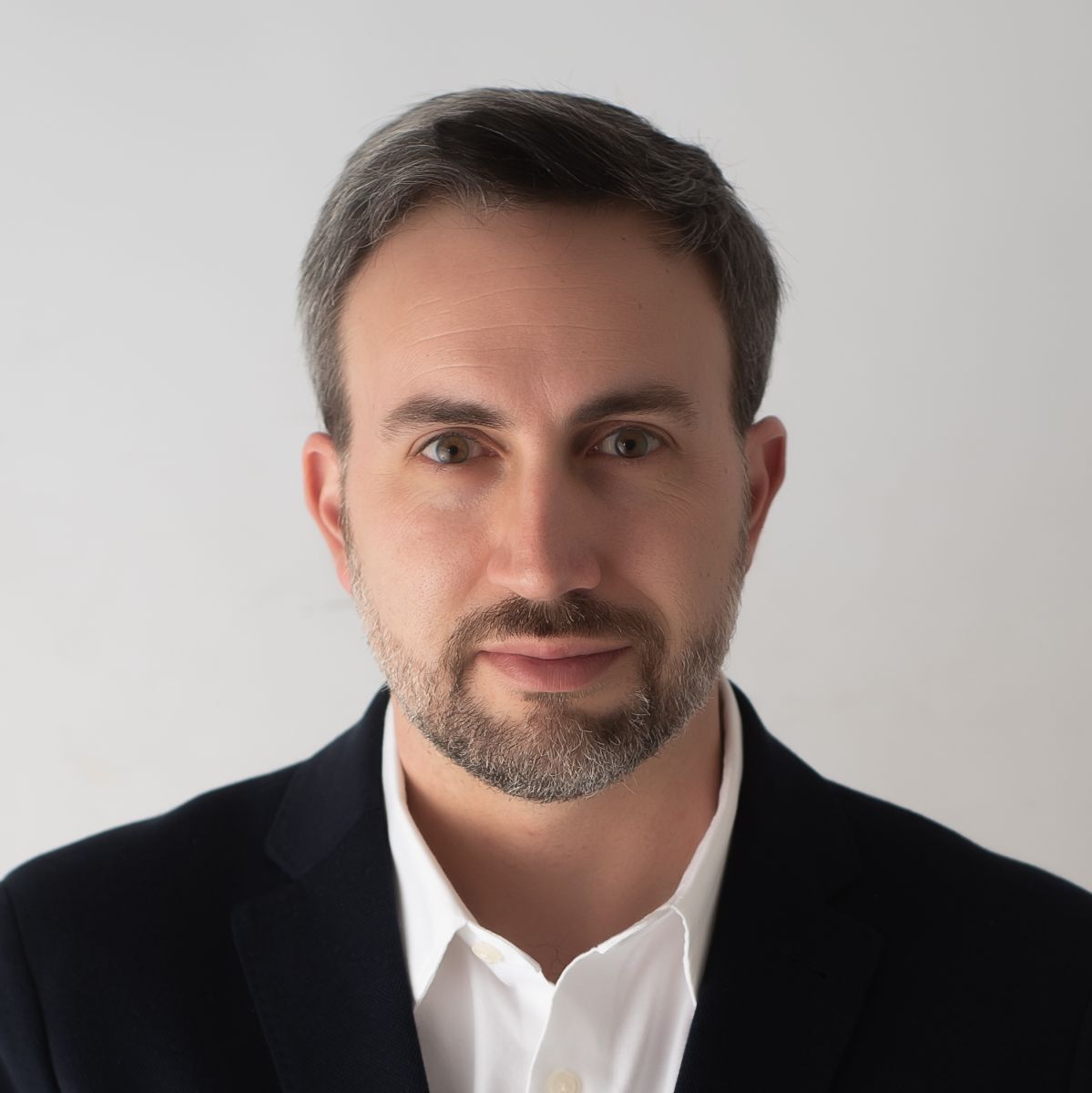 Light-skinned male-presenting person with dark hair wearing a black jacket with white button-up shirt in front of a light gray background