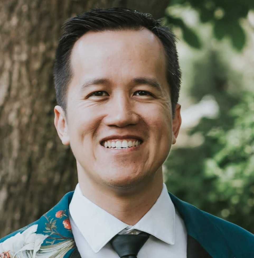 A smiling male-presenting individual of Asian heritage with dark hair wearing a green, floral-print jacket and tie against an outdoor background