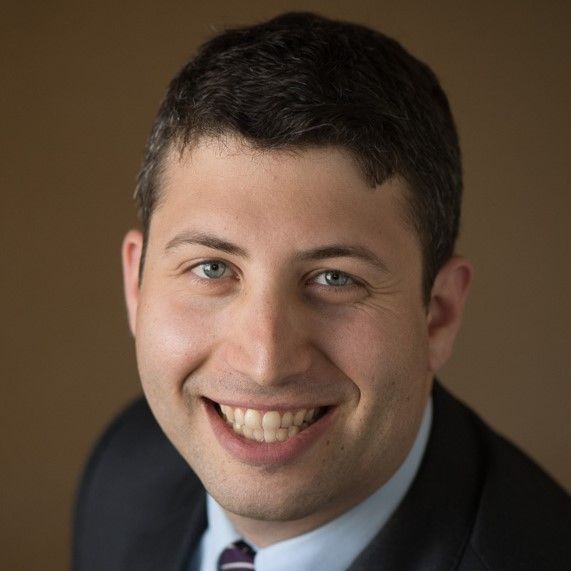 Smiling light-skinned, male-presenting individual with short dark hair wearing a black jacket on a brown backdrop.