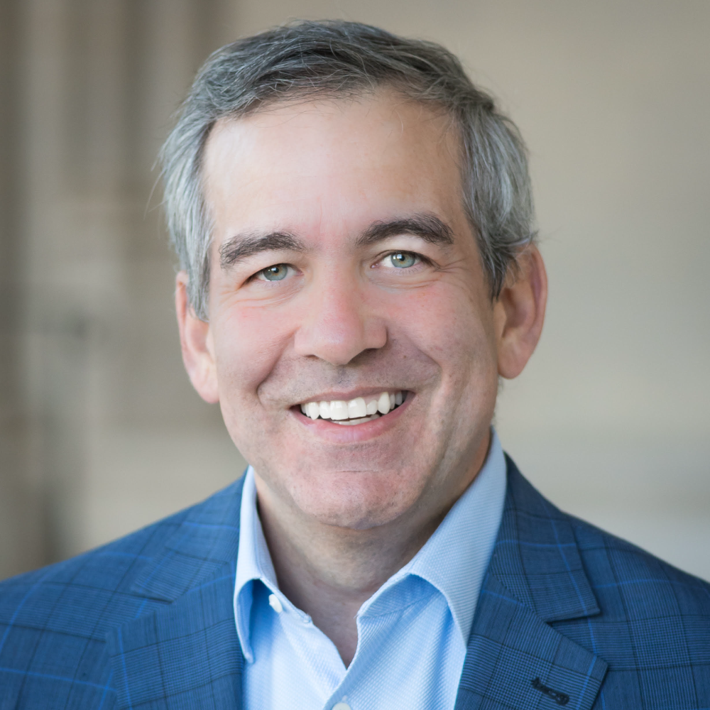 A smiling light-skinned male-presenting person wearing a blue suit jacket and button-up shirt with a blurred gray background