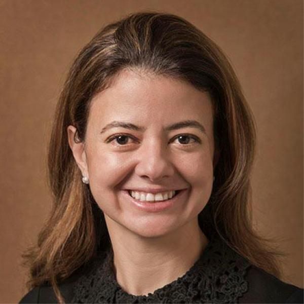 A smiling light-skinned female-presenting person with dark brown hair wearing a black shirt against a brown gradient background