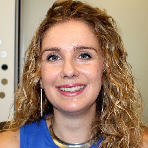 A smiling light-skinned female-presenting person with blonde hair wearing a blue shirt against a light background