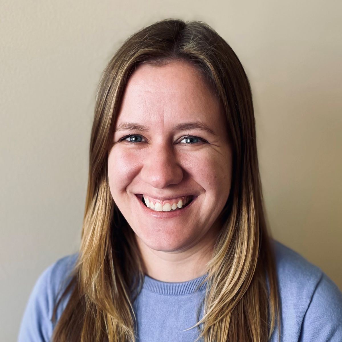 Smiling light-skinned, female-presenting individual with long length blonde hair wearing a grey shirt on a beige backdrop.