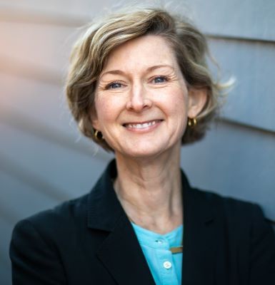 Smiling, female-presenting individual with blonde hair in a dark suit in front of a gray background