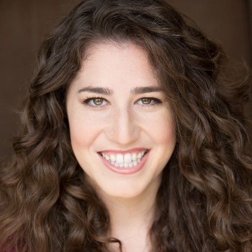 Smiling light-skinned, female-presenting individual with long dark hair wearing a dark shirt on a brown backdrop.