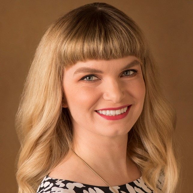 Smiling light-skinned, female-presenting individual with long blonde hair wearing a black and white shirt on a grey backdrop.