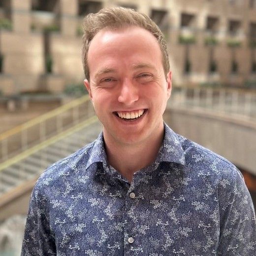 A smiling light-skinned male-presenting individual wearing a patterned blue shirt with a blurred background