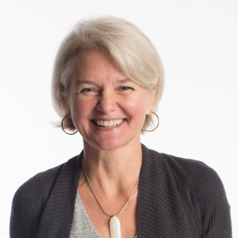 A smiling light-skinned, female-presenting with white hair wearing a dark sweater over a light gray shirt against a light background