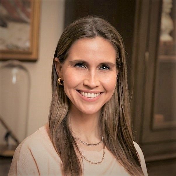 Smiling light-skinned, female-presenting individual with long dark hair wearing a beige shirt on a indoor backdrop.