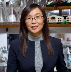 A smiling female-presenting individual of Asian heritage wearing a navy suit jacket over a gray sweater against a blurred background