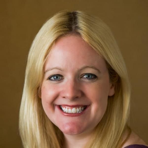 Smiling light-skinned, female-presenting individual with long blonde hair wearing a black shirt on a brown backdrop.