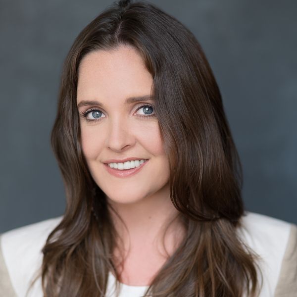 A smiling Caucasian female-presenting individual with dark brown hair wearing a white shirt against a dark gray background