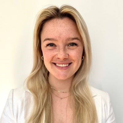 Smiling light-skinned, female-presenting individual with long blonde hair wearing a white jacket on a light backdrop.