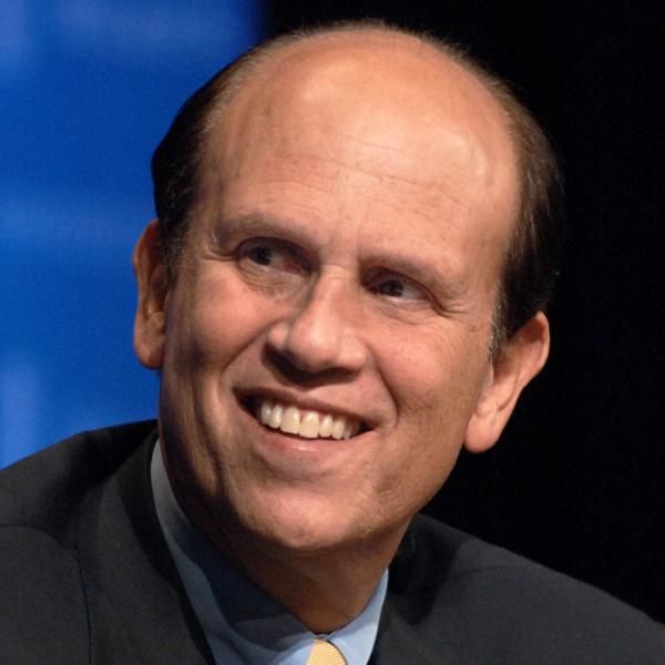 A smiling caucasian male-presenting individual wearing a dark suit and tie against a bright blue and black background