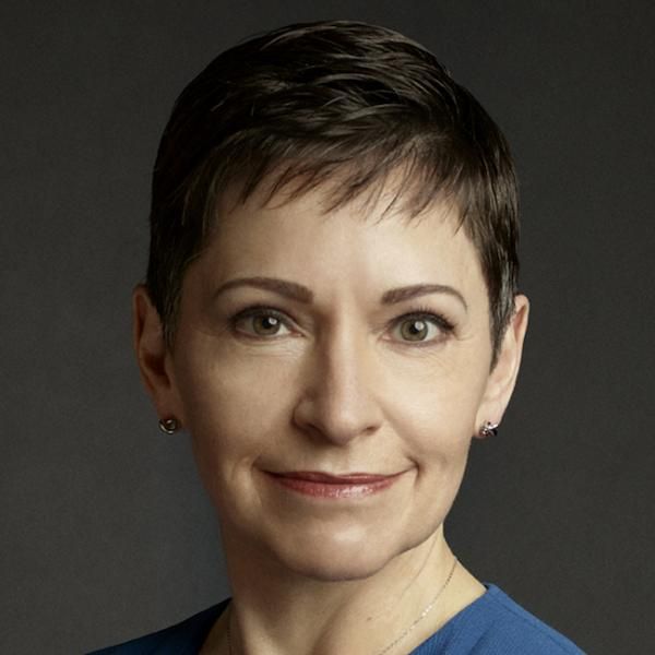 A female-presenting person with short dark hair wearing a blue top against a dark background.