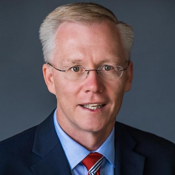 A smiling light-skinned male-presenting person wearing a navy blue suit and tie with gray hair and glasses against a gray gradient background