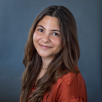 Smiling light-skinned, female-presenting individual with long dark hair wearing a red shirt on a grey backdrop.