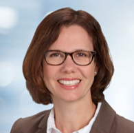 A smiling light-skinned female-presenting person with dark brown hair wearing glasses and a brown suit jacket over a white button-up shirt against a blue gradient background