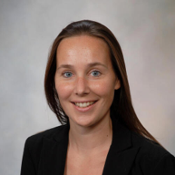 A smiling female-presenting light-skinned person with dark brown hair wearing a black suit jacket against a light gray background