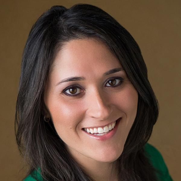 Smiling light-skinned, female-presenting individual with long dark hair wearing a green shirt on a brown backdrop.