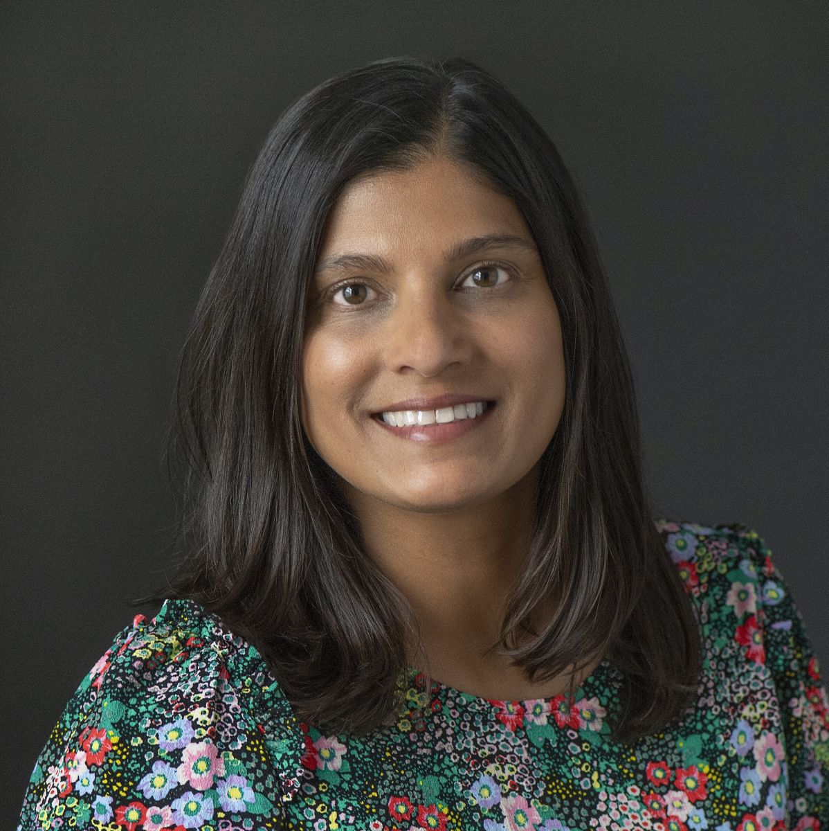 Smiling medium-skinned, female-presenting individual with medium length dark hair wearing a multi-colored shirt on a dark backdrop.