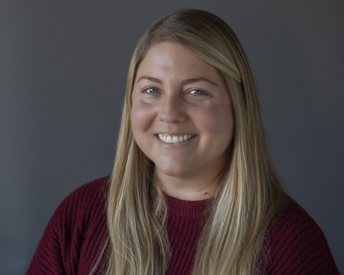 Smiling light-skinned, female-presenting individual with long length blonde hair wearing a burgundy shirt on a grey backdrop.